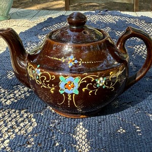 May include: A brown ceramic teapot with a lid and handle. The teapot is decorated with colorful floral designs in blue, yellow, and green. The teapot is sitting on a blue patterned surface.