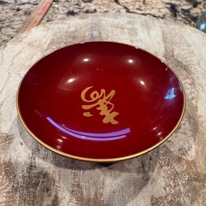 May include: A small, red lacquer bowl with a gold rim and a gold kanji character in the center.