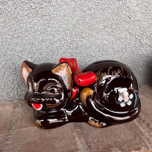 May include: A brown ceramic cat piggy bank with a red bow and a white flower on its side. The cat is lying down with its head turned to the side. The piggy bank has the words "The Curley" written on its side.