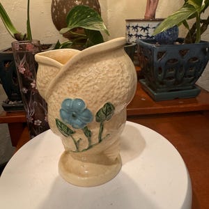 May include: A beige ceramic vase with a textured surface and a flared top. The vase features a blue flower design with green leaves. The vase is on a white surface, with other potted plants in the background.
