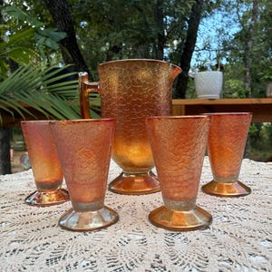 Puede incluir: Un juego vintage de jarra y cuatro vasos de vidrio ámbar. El conjunto presenta un diseño texturizado y agrietado. La jarra tiene un asa y un pico. Los vasos tienen una base de pedestal. El conjunto se exhibe sobre un mantel de ganchillo color crema.