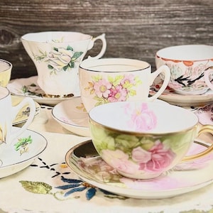 May include: A collection of six vintage teacups and saucers with floral patterns. The teacups are in various shades of pink, white, and yellow. The saucers are decorated with delicate floral designs.