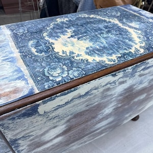 Puede incluir: Una mesa plegable vintage con un acabado envejecido. La tapa presenta un diseño floral y paisajístico en azul y blanco. Los bordes son de madera oscura y los lados tienen un aspecto desgastado y envejecido. La mesa tiene patas pequeñas y redondeadas.