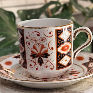 May include: A white teacup and saucer set with a decorative floral and geometric pattern in black, orange, and gold. The cup and saucer have gold trim. The design includes a bow and diamond shapes.