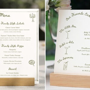May include: Two wooden menu stands with white paper inserts. The left stand has a menu for family style salads, pizzas, and desserts. The right stand has a menu for cocktails, including "The Little Dan", "Ralph-a-Rita", and "The Sycamore".