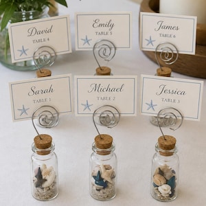 May include: Six clear glass bottles filled with sand and small seashells, each topped with a cork and a silver wire spiral holder. Each holder displays a rectangular white place card with a blue border, a blue starfish icon, and names like David, Emily, James, Sarah, Michael, and Jessica with table numbers.