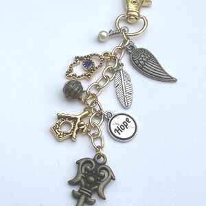 May include: A gold-toned keychain with multiple charms, including a key, feather, wing, and a charm with the word "Hope." Additional charms include a blue-stoned hamsa hand and a decorative bead. The keychain has a lobster clasp.