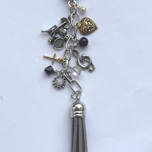 May include: Silver-toned keychain with various charms, including a gold heart, cross, musical note, and a gray tassel. The charms are connected by a chain and a clasp at the top.