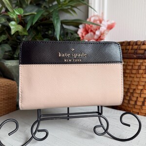 May include: A black and pale pink Kate Spade wallet with gold lettering, displayed on a decorative black metal stand. The wallet is two-toned, with a black top section and a pale pink bottom section.