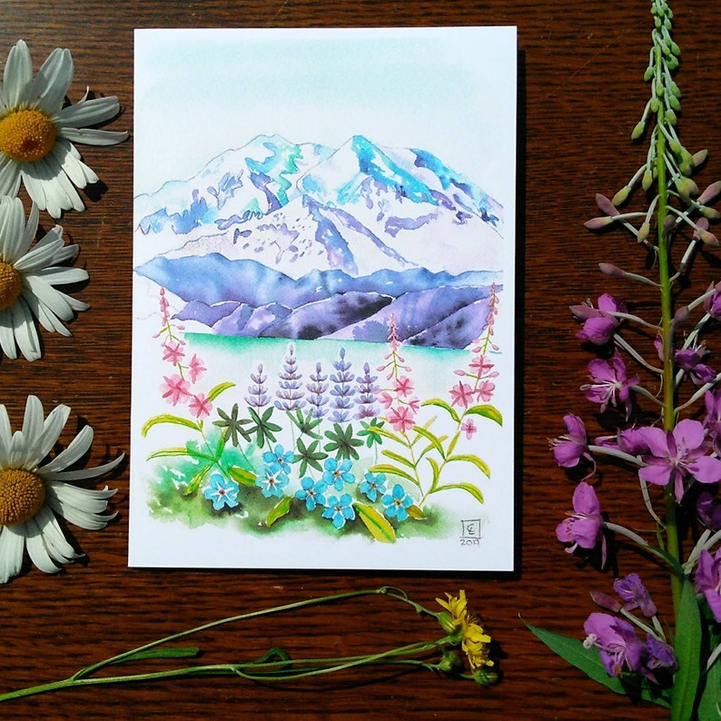 May include: Watercolor painting of a mountain landscape with snow-capped peaks, a lake, and colorful wildflowers. The artwork features shades of blue, purple, green, and pink. Daisies and other flowers are arranged around the painting on a wooden surface.