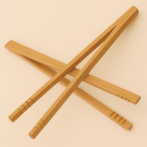 Puede incluir: Pinzas de bambú de madera con acabado natural. Las pinzas tienen forma rectangular con un extremo de agarre ranurado. Las pinzas están diseñadas para servir alimentos. El fondo es de color beige claro.