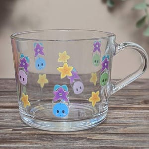 May include: A clear glass mug featuring cartoon characters and yellow stars. The mug has a handle and is on a wooden surface. The design includes purple, blue, and green characters. The mug is suitable for hot or cold drinks.