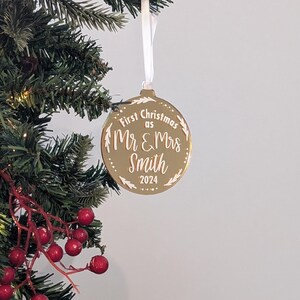 May include: A gold-coloured Christmas bauble with the text "First Christmas as Mr & Mrs Smith 2024" in white script. The round bauble has a white ribbon and is displayed on a Christmas tree.