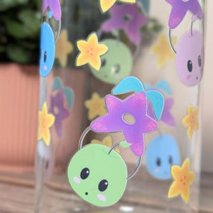 May include: A clear glass water bottle adorned with cartoon illustrations. The design includes stars, green apple-like characters, and other whimsical shapes in purple, yellow, blue, and green. The bottle is transparent.