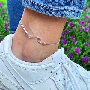Puede incluir: Una tobillera de tono plateado con un dije central en forma de ola unido a un cordón fino de color claro. La tobillera se lleva alrededor del tobillo, colocada sobre una zapatilla deportiva blanca y bajo el dobladillo de unos vaqueros azules.