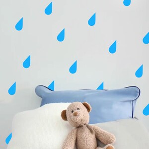 Op de afbeelding: Een wit bed met een blauw kussen en een teddybeer. De muur achter het bed is versierd met blauwe regendruppel stickers.