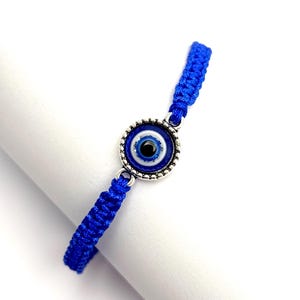 May include: A blue woven bracelet featuring a silver-toned charm with a blue and white eye design. The bracelet is made of a woven blue cord. The eye charm has a black pupil and is surrounded by a silver-colored metal.