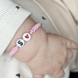 Może przedstawiać: Różowa bransoletka z makramy z białymi koralikami. Jeden koralik ma czarną literę "S", a drugi czerwone serce. Bransoletka jest na nadgarstku dziecka. Tło jest w miękkim, neutralnym kolorze.
