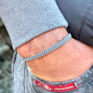 May include: A light gray macrame bracelet is worn on a wrist. The bracelet is made of woven cord and has a simple, minimalist design. The person is wearing a gray long-sleeve shirt and gray jeans.