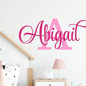 Può includere: Adesivo da parete rosa e bianco con il nome "Abigail" in un font corsivo. La prima lettera "A" è più grande e contornata di rosa.