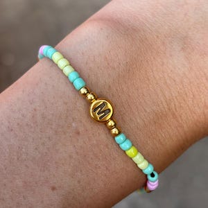 Puede incluir: Una pulsera de cuentas con un patrón repetitivo de pequeñas cuentas de semillas en tonos pastel rosa, amarillo y turquesa. El centro de la pulsera muestra una cuenta redonda dorada con una letra M negra impresa, flanqueada por cuentas doradas.