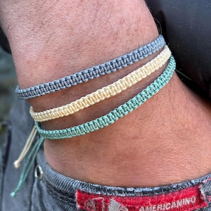 Puede incluir: Tres pulseras de macramé en tonos gris, amarillo y verde azulado. Las pulseras se llevan en la muñeca, con los extremos de la pulsera verde azulado visibles. Las pulseras están hechas de cordón tejido y tienen un diseño sencillo y minimalista. Un accesorio informal.