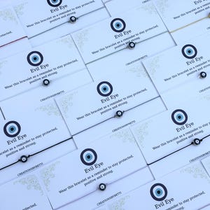 Könnte beinhalten: Mehrere dünne Schnurarmbänder mit einem kleinen runden Schutzaugen-Anhänger in Blau, Weiß und Schwarz. Jedes Armband ist auf einer weißen Karte befestigt, die mit dem Text Evil Eye und einem Hinweis zum Schutz bedruckt ist.
