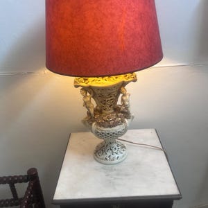 Op de afbeelding: Een tafellamp met een dieprode, getextureerde lampenkap en een crèmekleurige voet met ingewikkelde opengewerkte latwerkpatronen, bloemendetails en twee kleine gebeeldhouwde figuren aan weerszijden van het urnvormige lichaam.