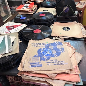 May include: A collection of black vinyl records stacked on a table, some housed in paper sleeves. One sleeve features blue text reading "ERST DURCH DEN ELECTROLA PLATTENSPIELER IST IHR EMPFANGSGERÄT VOLL AUSGENUTZT" alongside a graphic of a record player.