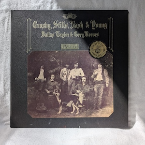 Może przedstawiać: Czarna okładka winylowego albumu z fotografią Crosby, Stills, Nash & Young z Dallas Taylorem i Gregiem Reevesem. Tytuł albumu to "Déjà Vu", a okładka zawiera złotą płytę.