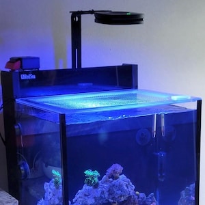 May include: A rectangular aquarium with a black frame and a built-in light fixture. The tank is filled with water, coral, and fish. The aquarium sits on a light-colored cabinet. The brand name "Red Sea" is visible on the top.