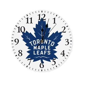 Peut inclure: Horloge blanche avec un motif de feuille d'érable bleue et les mots "Toronto Maple Leafs". Le cadran de l'horloge a des chiffres et des aiguilles noirs.
