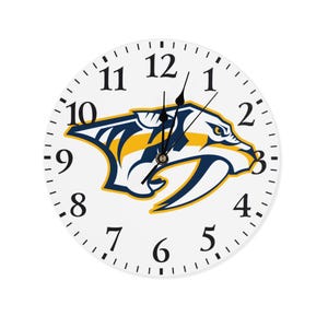 May include: White wall clock featuring the Nashville Predators logo in navy blue, gold, and white. The clock face has black numerals and minute markers. The clock hands are black.