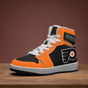 Puede incluir: Una zapatilla de caña alta con un diseño en negro y naranja, cordones blancos y suela blanca. El lateral muestra el logotipo de los Philadelphia Flyers en blanco y naranja, con el texto Philadelphia Flyers impreso en el cuello naranja superior.