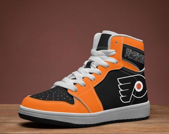 Pode incluir: Um tênis de cano alto com um esquema de cores preto e laranja, cadarços brancos e sola branca. A lateral exibe o logotipo do Philadelphia Flyers em branco e laranja, com o texto Philadelphia Flyers impresso na gola laranja superior.