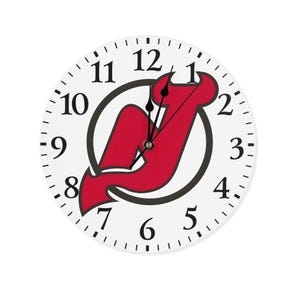 May include: A round white wall clock featuring the New Jersey Devils logo in red and black. The clock face has black numerals and minute markers. The hands are black, and the logo is centered.