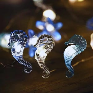 May include: Three blue, gray, and white seahorse-shaped string lights. The lights are strung on a clear wire and are illuminated with white LED lights.