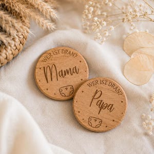 Könnte beinhalten: Zwei runde Holzscheiben mit eingraviertem Text und Grafiken. Eine Scheibe zeigt "WER IST DRAN?" über dem Wort "Mama" und einem Windelsymbol. Die zweite Scheibe zeigt "WER IST DRAN?" über dem Wort "Papa" und einem Windelsymbol mit kleinen Herzen.