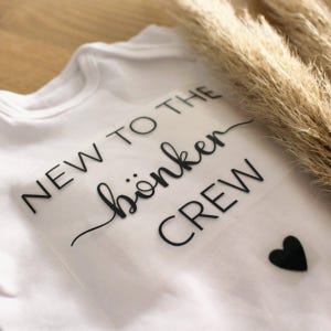 Könnte beinhalten: Weißer Baby-Body mit dem Text "NEW TO THE böken CREW" in schwarzer Schrift. Ein kleines schwarzes Herz befindet sich neben dem Text. Der Body ist aus weichem Material.