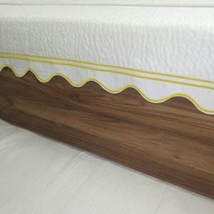 May include: A white bedspread with a scalloped edge and two yellow stripes. The bedspread is on a wooden bed frame.
