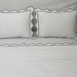 May include: White bed linen with a scalloped edge and black trim. The linen is folded and arranged on a bed.