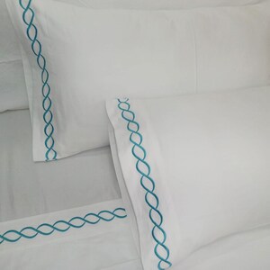 May include: White pillowcases and a fitted sheet with a teal chain stitch embroidery border. The bedding set is made of a smooth, crisp fabric. The design is simple and elegant.