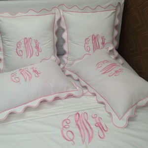 Puede incluir: Cuatro fundas de almohada blancas con bordes festoneados rosas e iniciales bordadas en rosa. Las fundas de almohada están dispuestas sobre una colcha blanca.