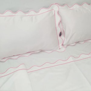 Op de afbeelding: Wit beddengoed met een roze gegolfde rand. Het beddengoed omvat een plat laken, een hoeslaken en twee kussenslopen.