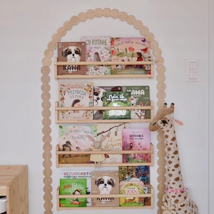 Op de afbeelding: Een houten boekenplank met meerdere planken, met kleurrijke kinderboeken. De boekenplank heeft de vorm van een boog en is versierd met een geschulpte rand. Een knuffelgiraf en een knuffeldier staan naast de boekenplank.