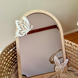 Op de afbeelding: Een boogvormige spiegel met een houten frame en een reflecterend oppervlak. Twee witte vlinderuitsparingen sieren het frame. De spiegel ligt in een rieten mand met een beige deken.