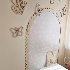 Op de afbeelding: Houten vlinder wanddecoratie in verschillende maten, samen met een frame met geschulpte rand en een interieur met madeliefjespatroon. Een houten letter "N" is ook aanwezig, met de naam "Nika" gegraveerd. De muur is geschilderd in een neutraal beige.