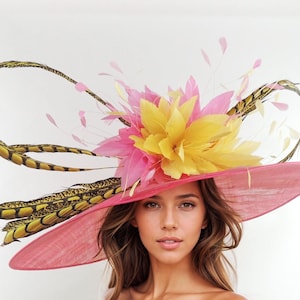 May include: A pink sun hat with a wide brim, adorned with yellow and pink floral accents and decorative feathers. The hat features a large, curved feather design in shades of gold and brown, creating a striking visual contrast.