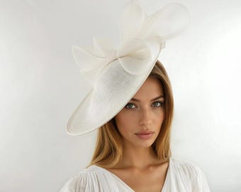 May include: An off-white fascinator hat featuring a wide, asymmetrical brim and a large, structured bow made of sheer, layered fabric loops. The hat is crafted from a textured material and sits at an angle on the head, creating a dramatic silhouette.