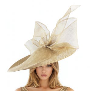 Puede incluir: Un sombrero grande, de color dorado, con ala ancha y lazo decorativo. El sombrero est&aacute; hecho de un material tejido y tiene un lazo transparente y aireado en la parte superior. Ideal para eventos formales.